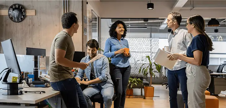 A group of five professionals standing and sitting around a workspace, engaged in a collaborative conversation.