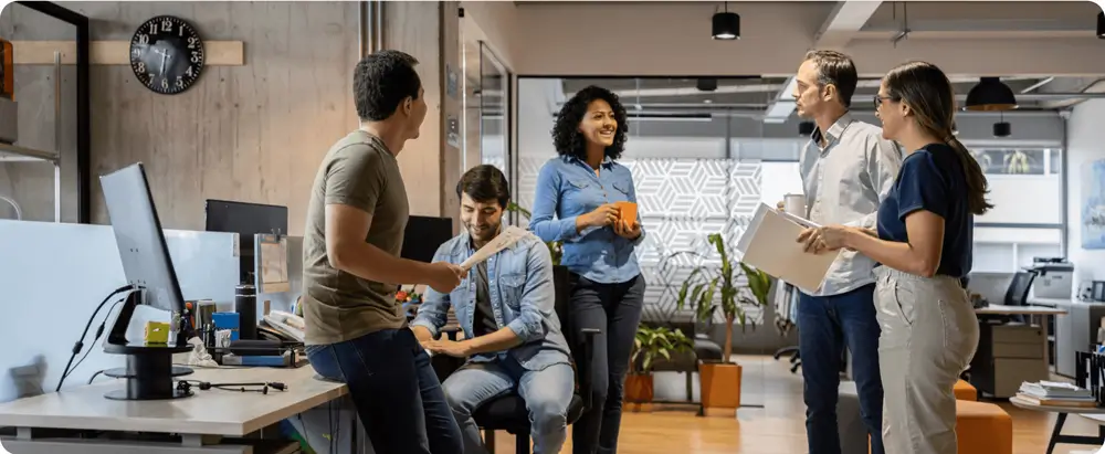 A group of five professionals standing and sitting around a workspace, engaged in a collaborative conversation.