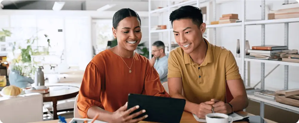 Two colleagues smiling and working together on a tablet in a bright, open office space, emphasizing teamwork and collaboration.