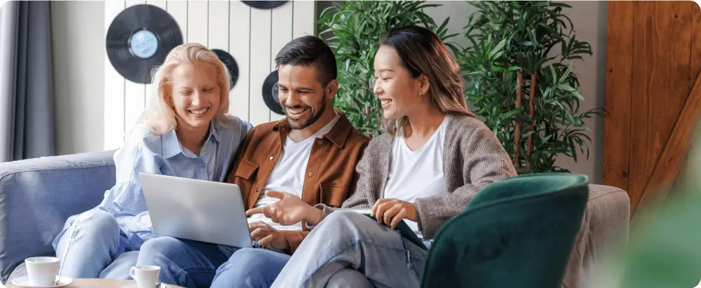 Three colleagues sitting on a sofa, smiling and collaborating on a laptop, in a casual, hybrid work environment.