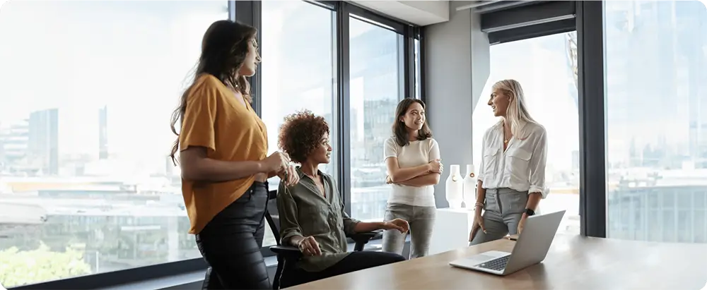 Four women conversing near a window in a modern office with cityscape view.