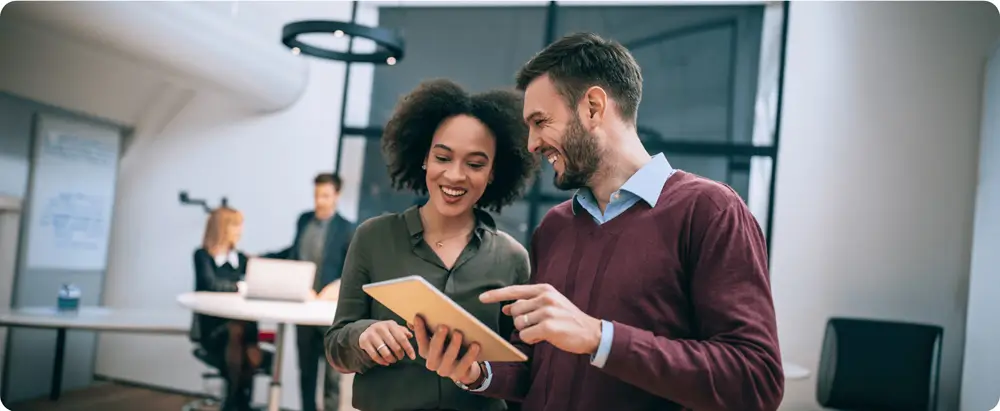 Two colleagues smiling and looking at a tablet in an office setting, with other team members working in the background