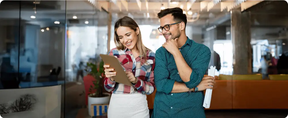 Two colleagues smiling and discussing something on a tablet in a modern office environment